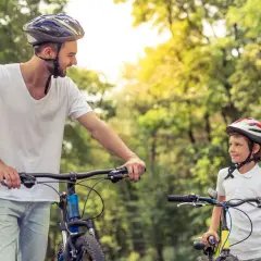 Ojciec i syn w kaskach rowerowych, patrzący na siebie z uśmiechem, otoczeni zielenią.