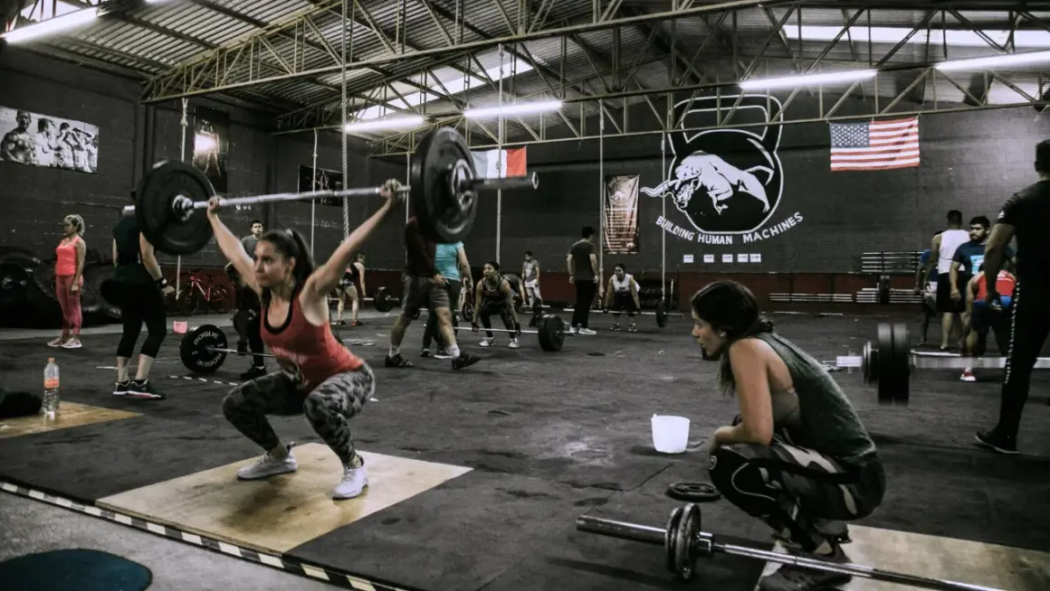Intensywny trening w stylu czym jest crossfit. Kobieta podnosi sztangę, inni ćwiczą w tle.