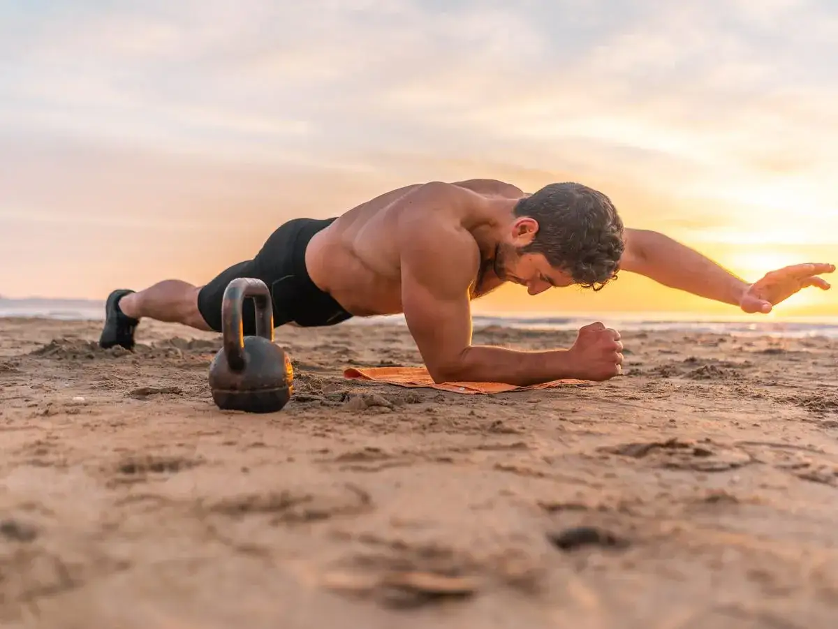 Mężczyzna na plaży wykonuje ćwiczenia na brzuch w deszczu. Kettlebell obok niego sugeruje intensywny trening.